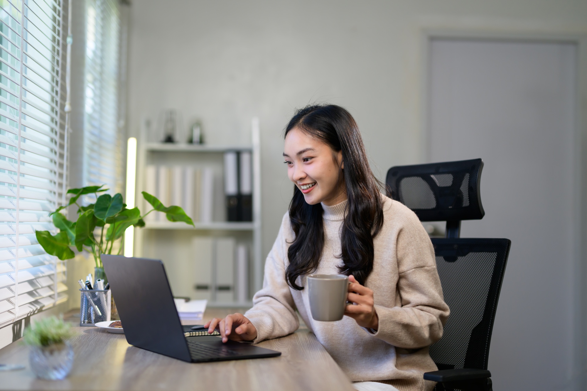 Young asian businesswoman sitting on an ergonomic chair and drinking coffee while working on a lapto