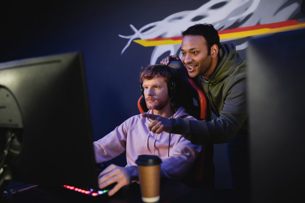 Positive indian man pointing with finger while friend playing video game on computer in gamming club